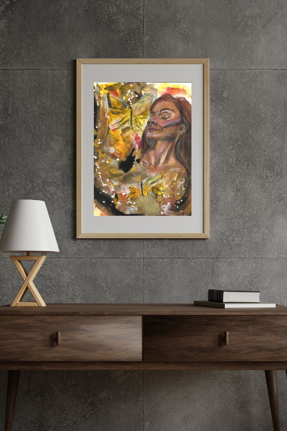 A framed acrylic painting titled A New Promise hanging on a dark textured wall above a wooden console table. The artwork features a serene woman with closed eyes, surrounded by golden butterflies, symbolizing transformation. The setting includes a minimalist lamp and stacked books, enhancing the modern and eclectic interior style.