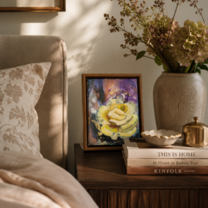 Small painting of a yellow rose on a wooden nightstand in a warm elegant bedroom